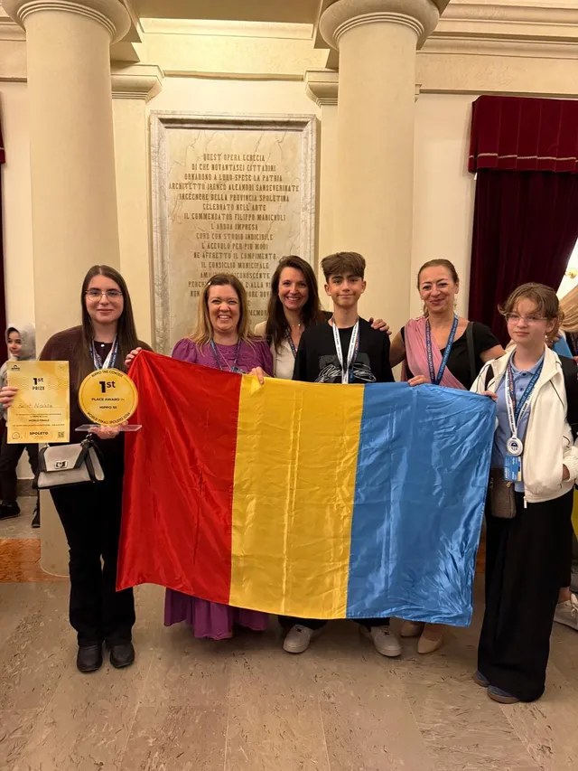 Șase persoane stau în rând în fața unui stâlp, ținând un steag tricolor și un trofeu, purtând medalii în jurul gâtului.
