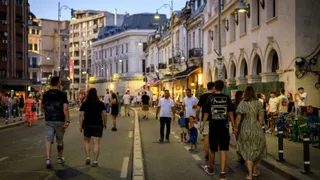 Oamenii merg pe o stradă cu asfalt, flancată de clădiri înalte și trotuare, sub lumina felinarelor de pe marginea drumului.