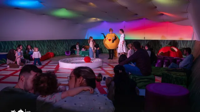 Oamenii stau pe pufuri colorate într-o încăpere iluminată în culori, privind un personaj costumat în formă de rotundă galbenă pe un fundal colorat.