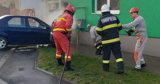 Patru bărbați îmbrăcați în uniforme de pompieri lucrează pe un teren verde lângă o mașină albastră și o clădire cu perete verde.