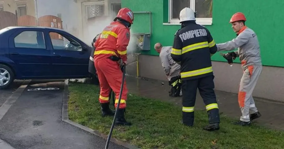 Patru bărbați îmbrăcați în uniforme de pompieri lucrează pe un teren verde lângă o mașină albastră și o clădire cu perete verde.