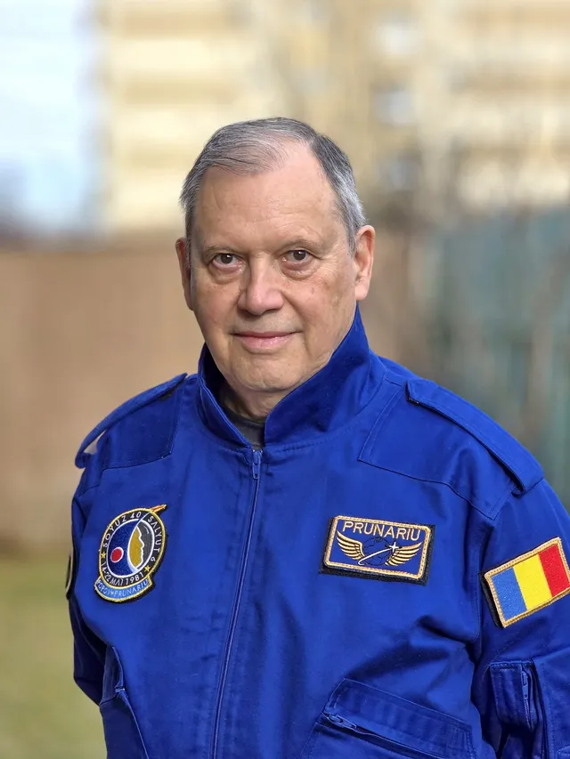 Un bărbat cu părul scurt și cămășă albastră poartă un costum de zbor albastru cu embleme pe piept și pe umăr.