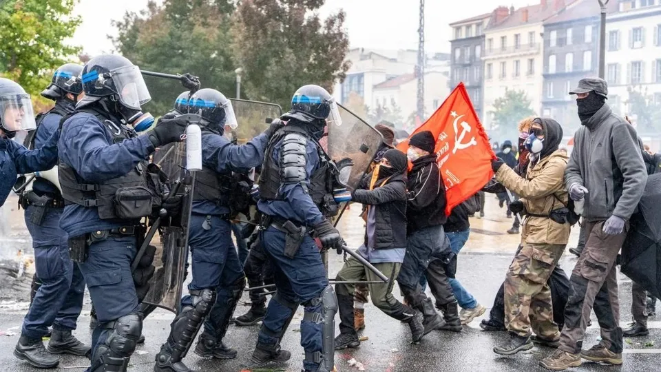 Polițiști în echipament de protecție și casci se confruntă cu un grup de persoane într-o zonă urbană cu un steag roșu ridicat.