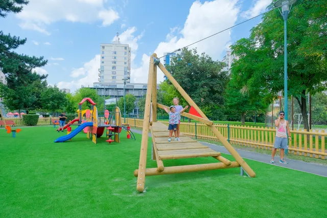 Un copil stă pe o structură de lemn, în timp ce un alt copil stă în picioare lângă ea, pe un teren cu iarbă artificială verde.