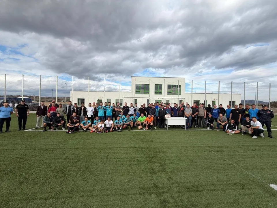 Un grup mare de bărbați stă poziționat pe un teren de fotbal cu iarbă artificială în fața unei clădiri albe sub un cer înnorat.