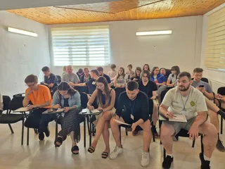 Un grup de persoane este așezat pe scaune într-o încăpere, unele dintre ele scriind pe foi de hârtie.