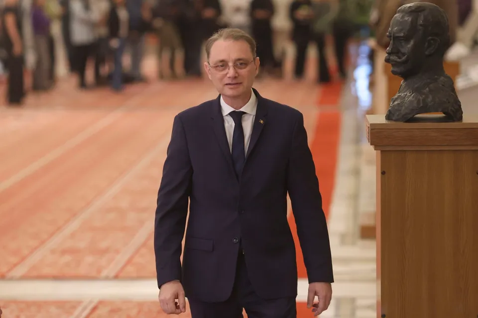 Un bărbat cu ochelari și costum închis la culoare stă în picioare într-o încăpere cu un bust pe un soclu în partea dreaptă.