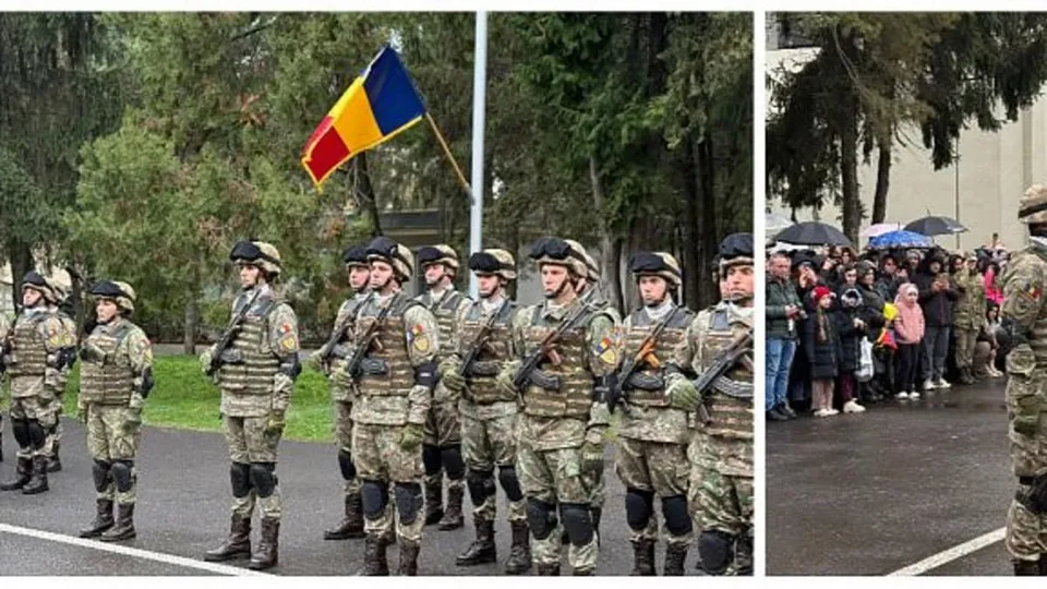 Soldați în uniformă de camuflaj și echipament de protecție stau în rând în fața unui steag și a unor copaci.