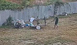 Două persoane se află pe un teren gol lângă un gard metalic, una împingând un cărucior cu obiecte și saci.