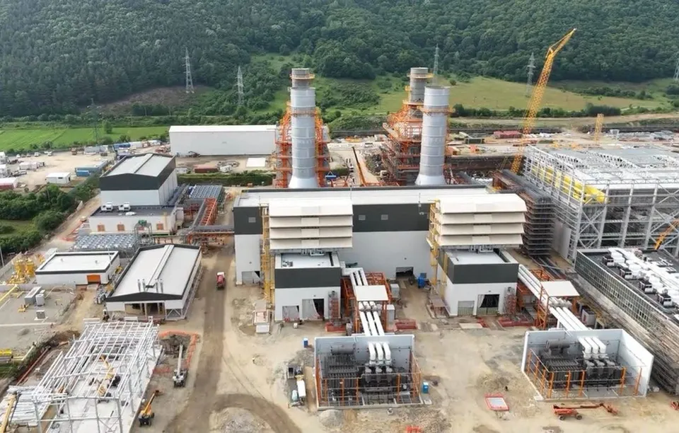 O vedere aeriană prezintă o centrală electrică în construcție cu două coșuri înalte, structuri metalice și utilaje de șantier pe un teren de pământ.