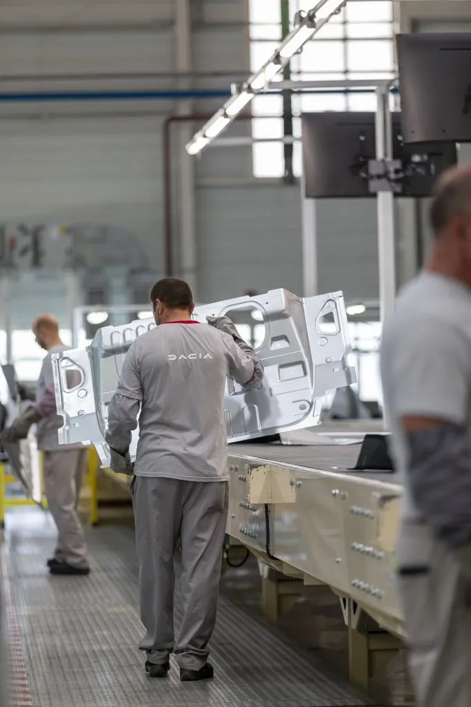 Un bărbat în haine de lucru gri manipulează o piesă metalică de caroserie pe o linie de asamblare într-o fabrică.