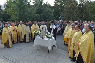 Preoți în veștminte liturgice aurii stau în cerc în jurul unei mese cu flori și un cristaler, într-o ceremonie religioasă la aer liber.