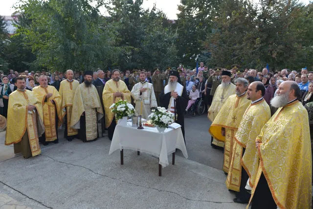 Preoți în veștminte liturgice aurii stau în cerc în jurul unei mese cu flori și un cristaler, într-o ceremonie religioasă la aer liber.