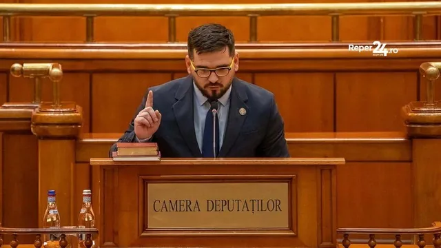 Un bărbat cu ochelari și costum în întunecire stă în spatele unui pupitru de lemn pe care scrie Camera Deputaților, ridicând un deget.
