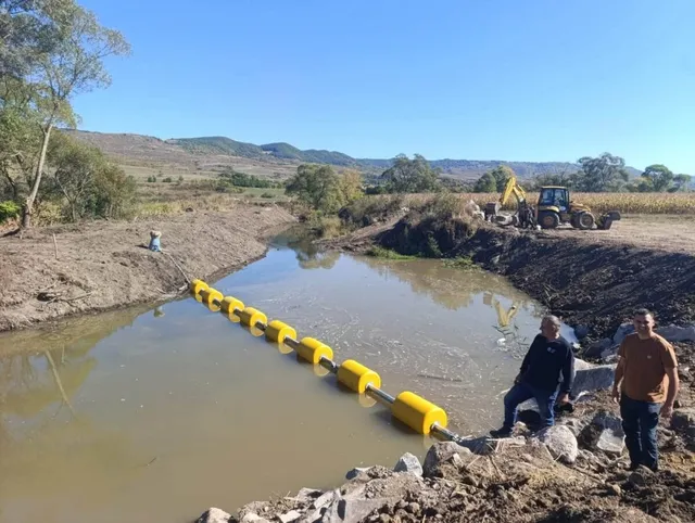 O barieră formată din cilindri galbeni plutind pe un curs de apă este instalată între două maluri de pământ.