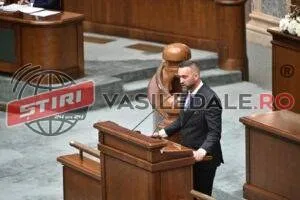 Un bărbat cu barbă, îmbrăcat în costum închis, stă în picioare în fața unui pupitru de lemn într-o încăpere cu mobilier din lemn.