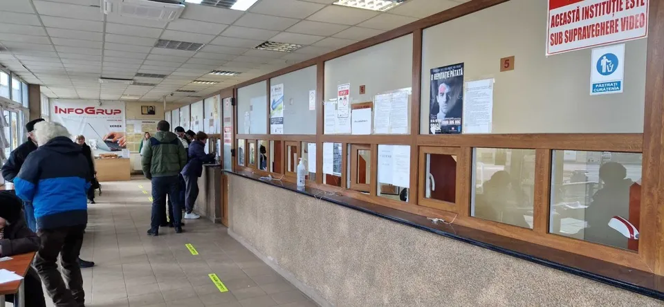 Oameni stau la coadă într-un spațiu public cu ghișee vitrate, panouri și afișe pe pereți.