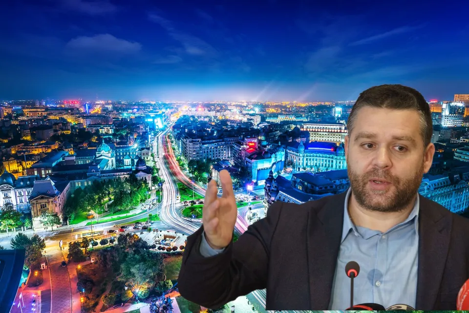 Un bărbat cu barbă, purtând sacou și cămașă, stă în prim-plan în fața unei panorame nocturne a unui oraș iluminat.