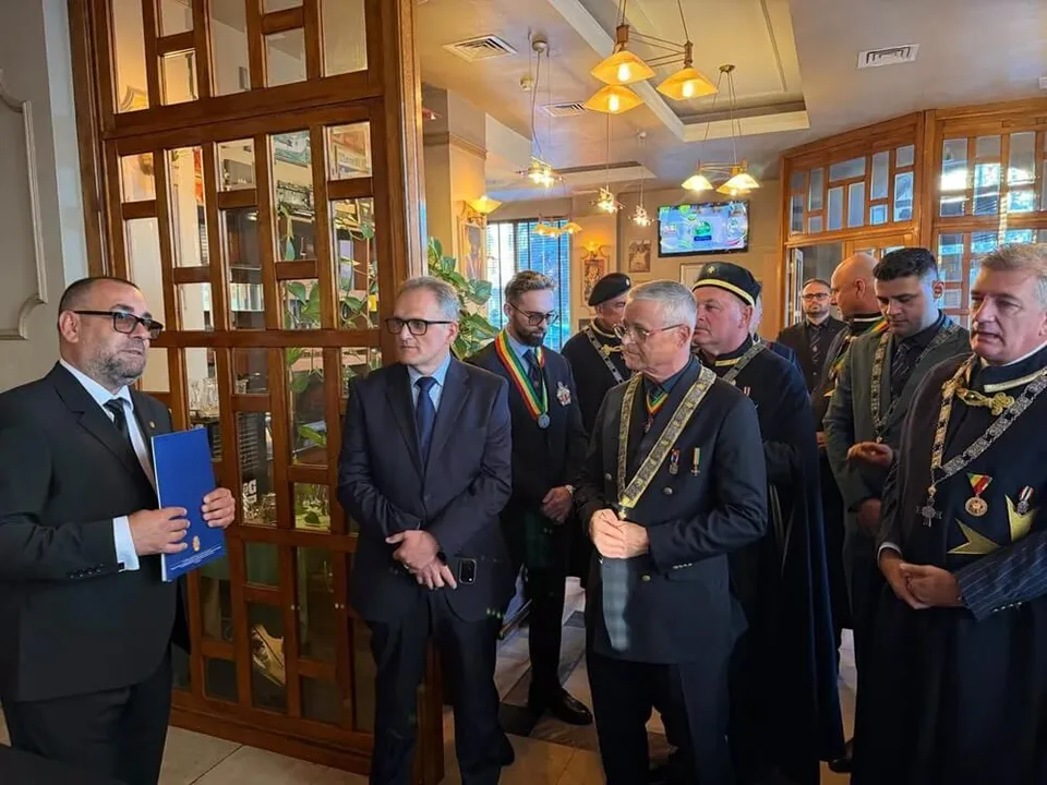 Un grup de bărbați în costume și uniforme oficiale cu panglici decorative stau în picioare într-o încăpere cu mobilier din lemn și corpuri de iluminat.