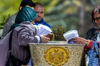 O persoană în jachetă mov și o alta cu ochelari ating un obiect metalic decorat cu motive aurii și pietre.