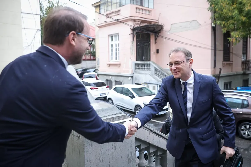 Un bărbat în costum în spatele căruia se află o clădire roz strângă mâna unui alt bărbat îmbrăcat în costum și cravată.