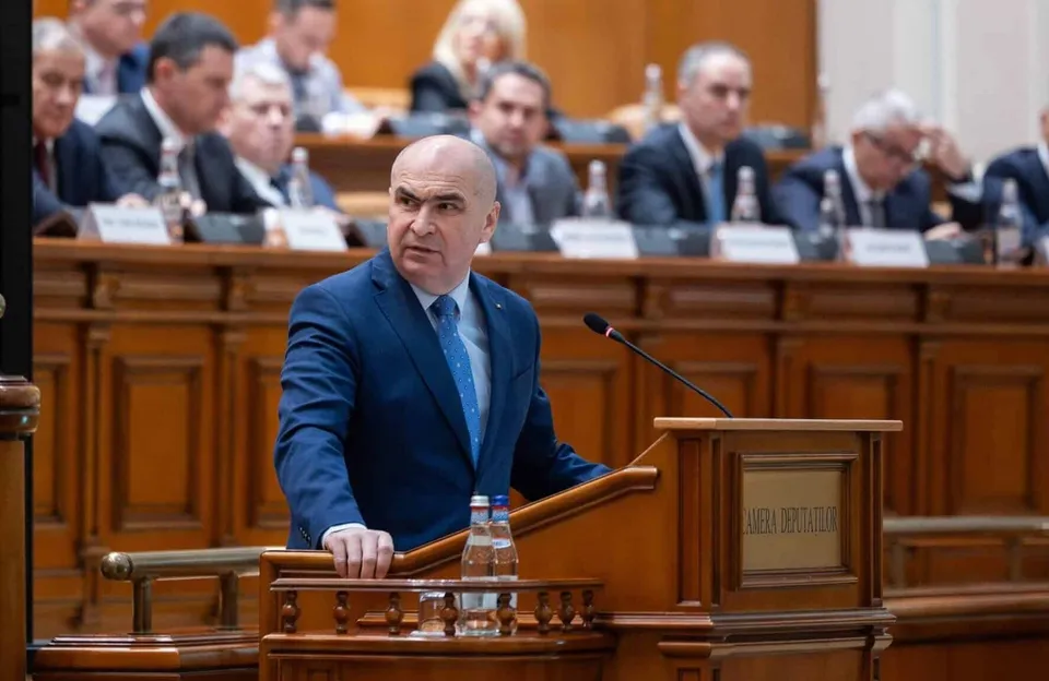 Un bărbat cu ochelari și costum albastru stă în spatele unui podium de lemn într-o sală cu mese lungi și oameni așezați în fundal.