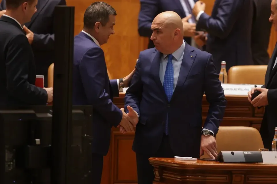 Un bărbat chel într-un costum albastru strânge mâna unui alt bărbat într-un costum albastru într-o încăpere cu mobilier din lemn.