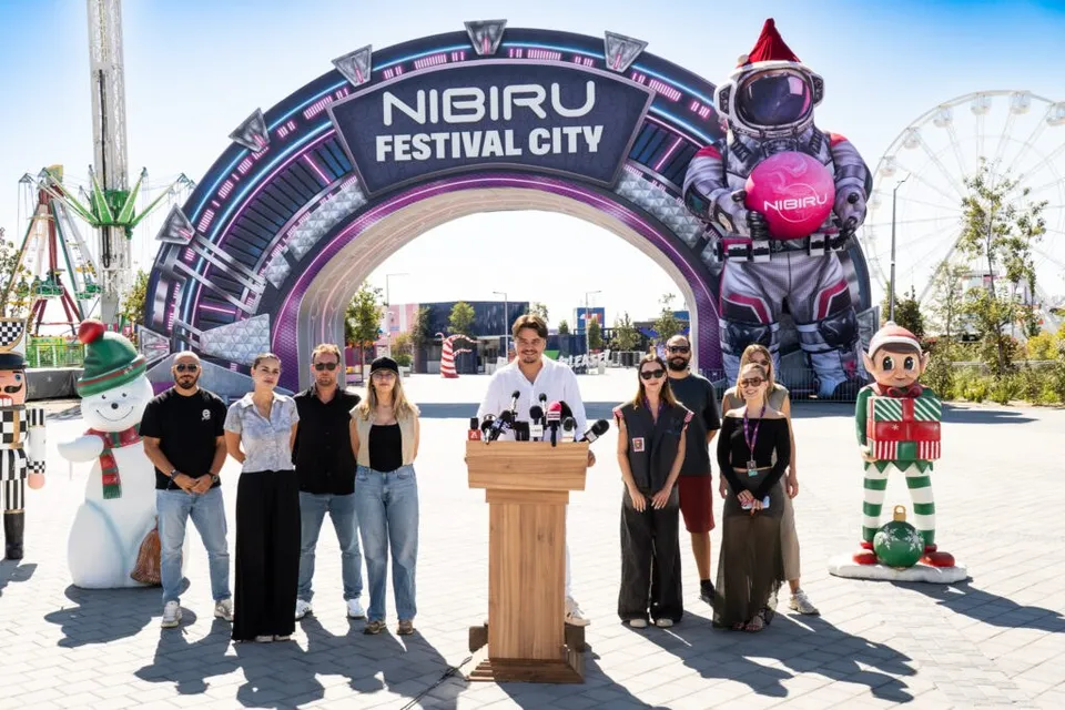 Un grup de persoane stă în fața unui arcul metalic pe care scrie NIBIRU FESTIVAL CITY, alături de figuri de talie mare.