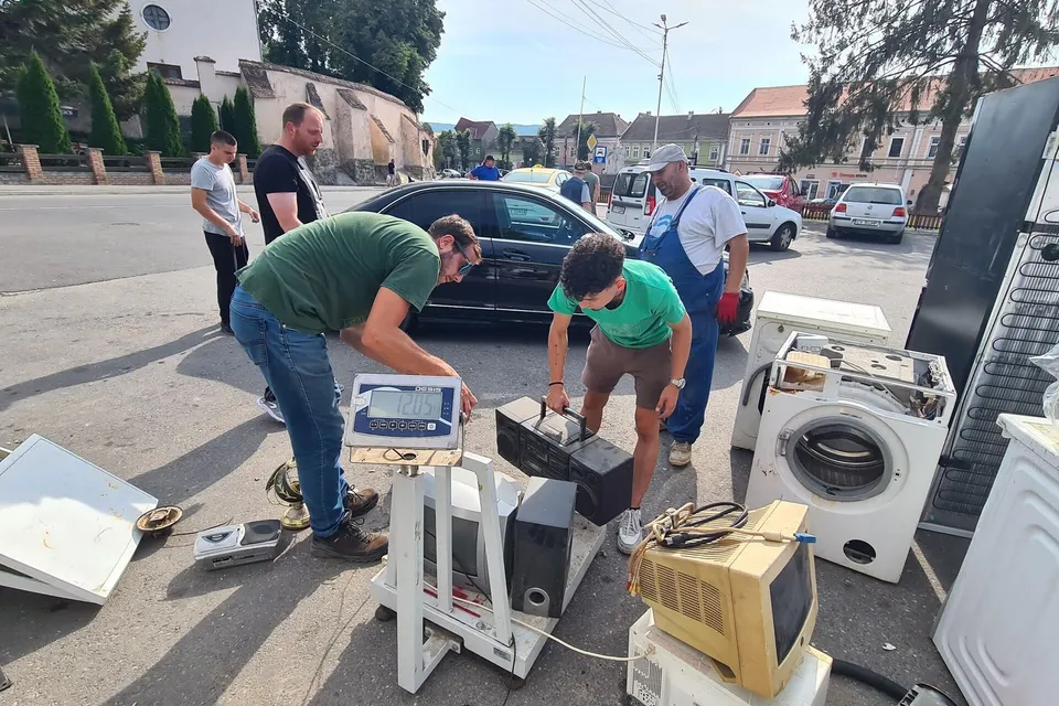 Un grup de bărbați stau pe o stradă lângă un cântar și diverse electrocasnice, inclusiv un monitor vechi și o mașină de spălat.