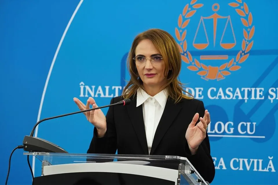 O femeie cu ochelari și sacou negru stă în spatele unui podium transparent în fața unui fundal albastru cu un simbol și text alb.