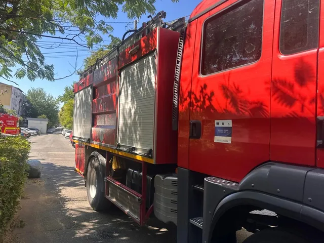 Un camion de pompieri roșu este parcat pe o stradă lângă un gard de vegetație sub un cer senin.