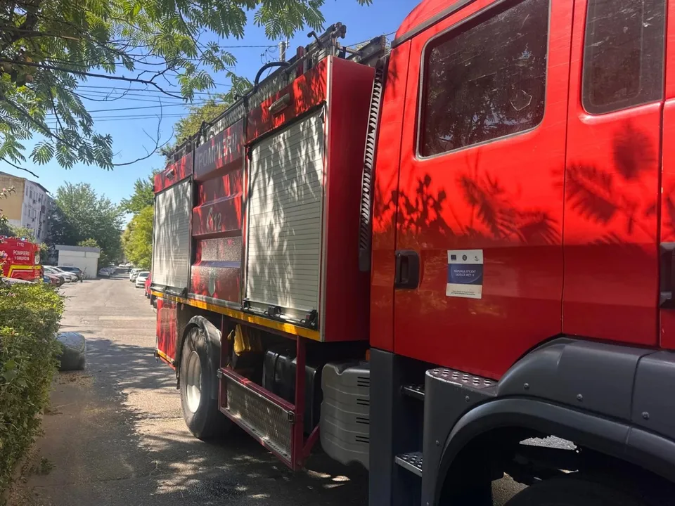 Un camion de pompieri roșu este parcat pe o stradă lângă un gard de vegetație sub un cer senin.