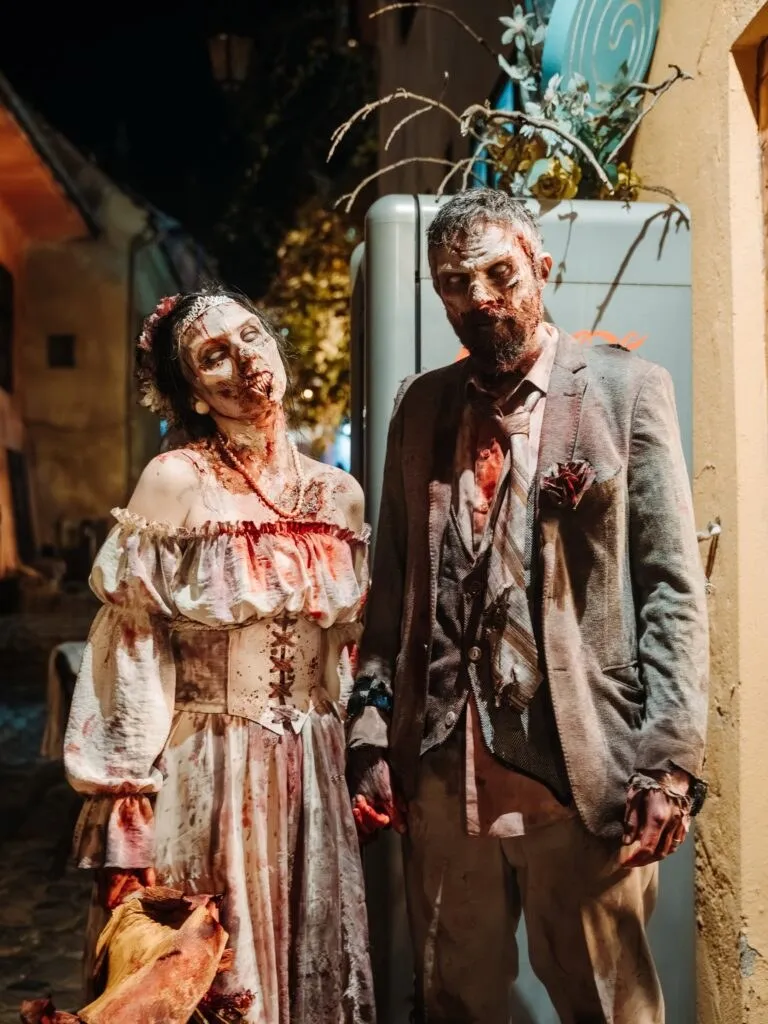 Două persoane îmbrăcate în costume de zombi, cu machiaj de efect horror și haine murdare, stau în picioare pe un drum noaptea.