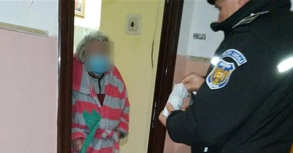 Un bărbat în uniformă neagră cu un emblema pe umăr ține un obiect alb în fața unei femei cu părul gri purtând un halat roz și alb.