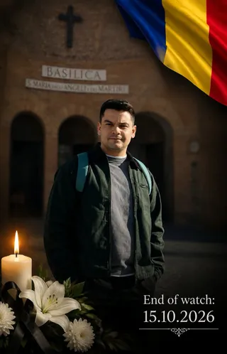 Un bărbat stă în fața unei clădiri cu o cruce pe perete, având un steag și flori cu o lumânare în prim-plan.