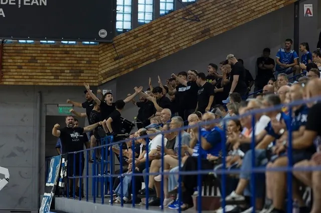Un grup de spectatori stă pe tribunele unei săli de sport, unii având brațele ridicate în sus.
