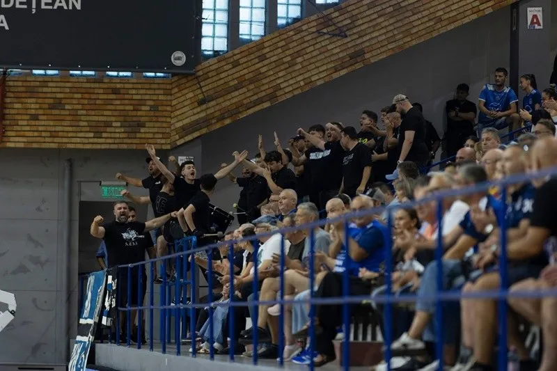 Un grup de spectatori stă pe tribunele unei săli de sport, unii având brațele ridicate în sus.