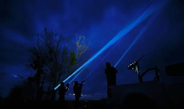 Siluete ale unor persoane în întuneric sub un cer albastru luminat de fascicule de lumină puternice și de ramurile unui copac.