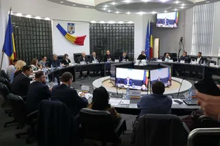 Oamenii stau la mese rotunde într-o sală de conferințe cu ecrane digitale, televizoare și steaguri pe pereți.