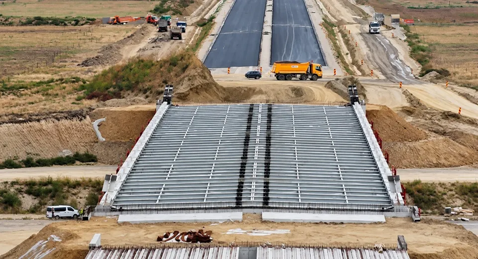 O structură de pod în construcție cu grinzi metalice gri este vizibilă în prim-plan, în fața unei porțiuni de drum asfaltat cu un camion galben.