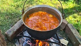 Un ceaun metalic cu mânere este așezat pe un suport de fier deasupra unui foc de lemne, conținând un preparat lichid de culoare portocalie.