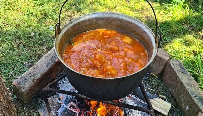 Un ceaun metalic cu mânere este așezat pe un suport de fier deasupra unui foc de lemne, conținând un preparat lichid de culoare portocalie.