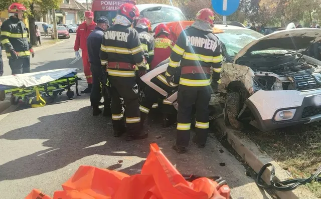 Mai mulți pompieri în echipament de protecție portocaliu și negru lucrează lângă un autoturism alb avariat pe un drum asfaltat.
