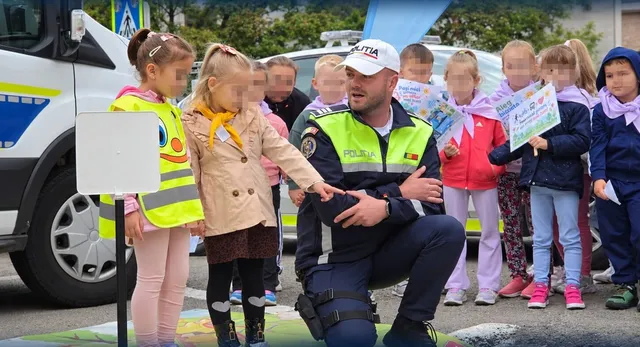 Un polițist cu uniformă și vestă reflectorizantă stă în genunchi în fața unui grup de copii pe o stradă lângă un vehicul alb.