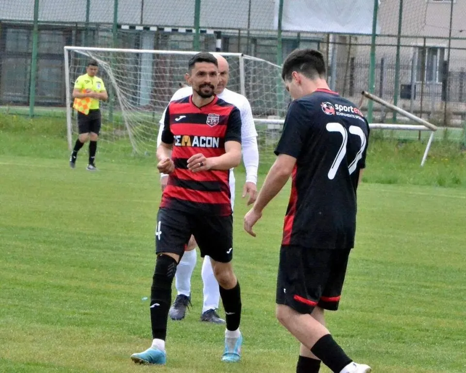 Un jucător de fotbal în echipament roșu și negru aleargă pe teren, în timp ce un arbitru și alți jucători sunt vizibili în fundal.