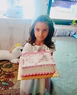 O copilă cu părul închis la culoare ține o prăjitură decorată cu roz și alb într-o cutie de carton pe un fundal de interior.