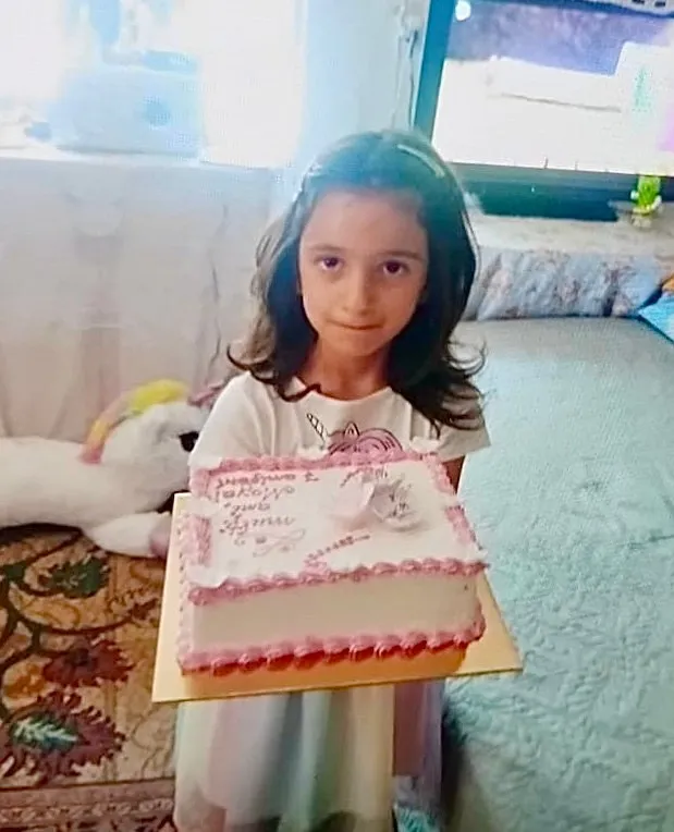 O copilă cu părul închis la culoare ține o prăjitură decorată cu roz și alb într-o cutie de carton pe un fundal de interior.