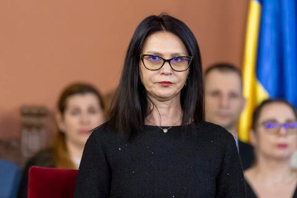 O femeie cu ochelari negri, păr lung și negru, îmbrăcată într-o bluză neagră, stă în picioare, privind direct spre cameră, într-o sală cu persoane în fundal.