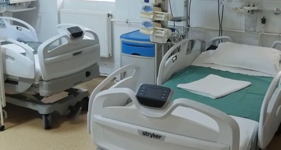 Două paturi de spital cu balustrade albe și lenjerie albă și verde sunt amplasate într-o cameră cu un radiator pe perete.