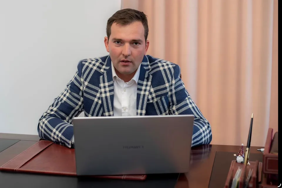 Un bărbat cu o jachetă albastră cu model de carouri albe stă la o masă în fața unui laptop deschis.